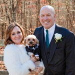 man and woman holding a small dog dressed in a tuxedo