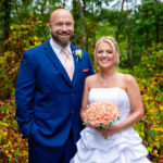 Bride in a strapless white gown holding a peach-colored bouquet and groom in a navy suit with a peach tie smiling together outdoors with greenery in the background.
