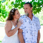 Woman in a white dress smiling and looking up at a man in a patterned shirt as they stand together outdoors under leafy green trees.