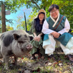 Two women in traditional and rustic-style clothing smiling and sitting outdoors next to a black-and-white pig