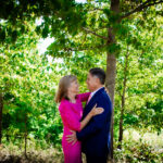 Woman in a bright pink dress smiling and embracing a man in a navy suit and pink tie as they stand together outdoors among green trees