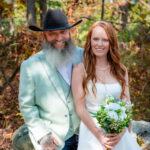 Bride in a white dress holding a bouquet of white flowers and groom in a light gray suit jacket with a black cowboy hat smiling together outdoors.