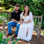 Couple sitting together on a wooden bench outdoors, holding hands, with the woman in a white dress and the man in a black shirt and jeans.