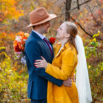 Bride and groom embracing in a colorful autumn forest, with the groom in a blue suit and tan hat holding a bouquet of vibrant flowers, and the bride in a yellow coat and veil smiling up at him amid the fall foliage.