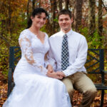 A woman in a white wedding dress sits next to a man wearing a shirt and tie on a bench outdoors in the woods