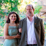 A woman and a man stand together outdoors, arm in arm, smiling warmly. The woman is wearing a light green sleeveless dress with subtle embroidered details, along with a necklace and bracelet. The man is dressed in a white button-down shirt and an olive-green jacket, accessorized with a necklace and a small heart-shaped pin. Behind them is a softly blurred background of greenery and fall colors, giving the scene a bright and pleasant atmosphere.
