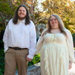 A man with long hair and a beard holds hands with a woman wearing a light yellow dress