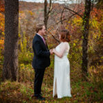 A man in a dark blue suit faces a woman dressed in a white wedding gown. They hold hands and look at one another.