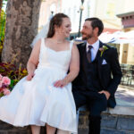 A woman in a white wedding dress sits next to a man dressed in a black suite. They are looking at one another and smiling.