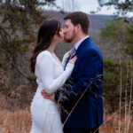A woman in a white dress kisses a man wearing a suit in a field