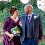A woman in a purple dress holds a boquet of flowers in one hand and the arm of a man wearing a blue suit in the other. Both look at each other and smile.