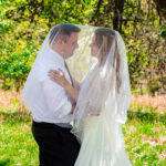 A newlywed couple stands face to face beneath a flowing white veil adorned with small pearl accents, sharing an intimate moment outdoors. The groom wears a white dress shirt with a gray tie and black trousers, while the bride wears a white wedding gown with long wavy blonde hair. They are surrounded by lush green trees and golden grass in a natural outdoor setting.