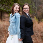 two women stand back to back and hold hands while smiling