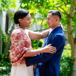 A bride and groom embrace face-to-face outdoors in front of a courthouse building, surrounded by green trees, shrubs, and ivy-covered ground. The bride has short dark hair and wears a red and white patterned kimono-style jacket over a cream dress, with one arm draped around the groom's shoulder. The groom has short dark hair and wears a navy blue blazer with a peacock feather boutonniere, light-colored pants, and a white shirt, with his arm around the bride's waist. Both are smiling and looking at each other. A small watermark logo appears in the bottom right corner.