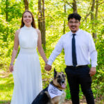 a bride and groom hold hands and smile facing forward with their german shepherd dog sitting in front of them